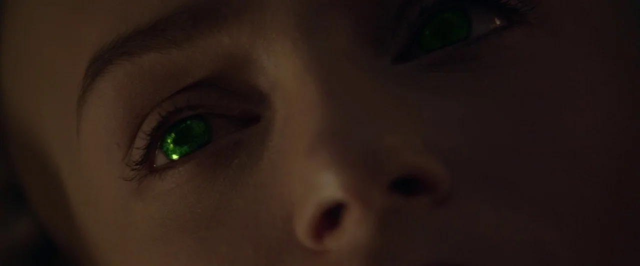 Starry Eyes (2014) movie still: closeup — A close-up of a human face in a dark, shadow-filled environment. The subject's eyes are glowing with a…