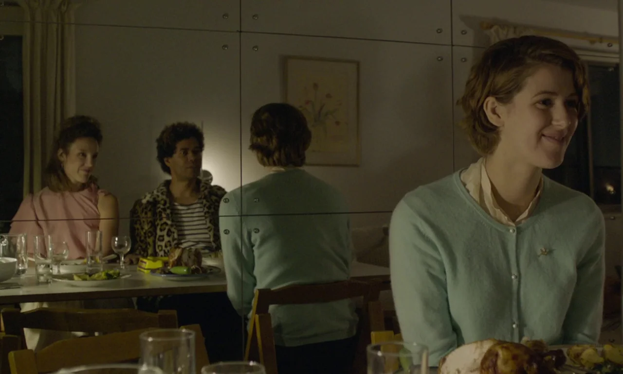 The Souvenir (2019) movie still: medium — A group of people sit at a table in front of a mirrored wall. The foreground shows a woman in a light blue…