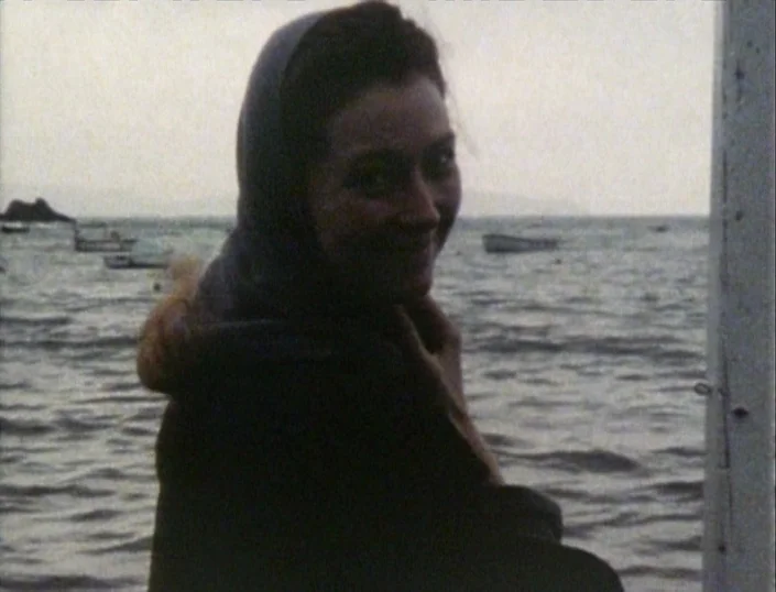 Glitterbug (1994) movie still: medium — A woman stands near a body of water under an overcast sky. Several small boats are visible floating in the…