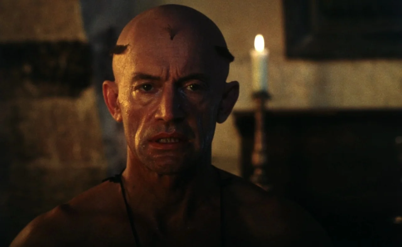 The Pit and The Pendulum (1991) movie still: closeup — A dimly lit room with rough stone walls and a single flickering candle. A bald middle-aged…