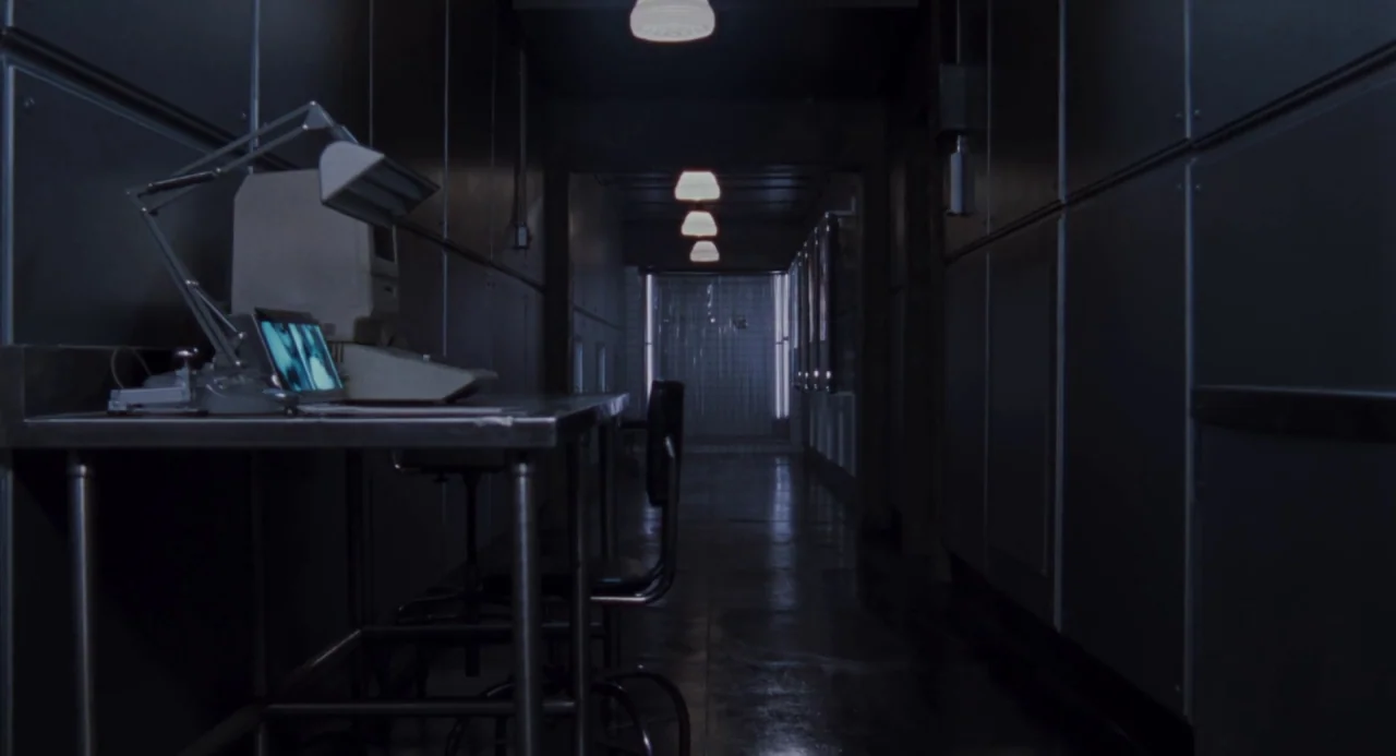 Martyrs (2008) movie still: wide — A dark, narrow corridor features grey metallic walls and a series of recessed ceiling lights. A metal desk with a…
