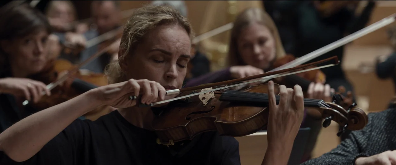 Tár (2022) movie still: closeup — A woman plays a violin within an orchestra, surrounded by other musicians. The backdrop consists of blurred wooden…
