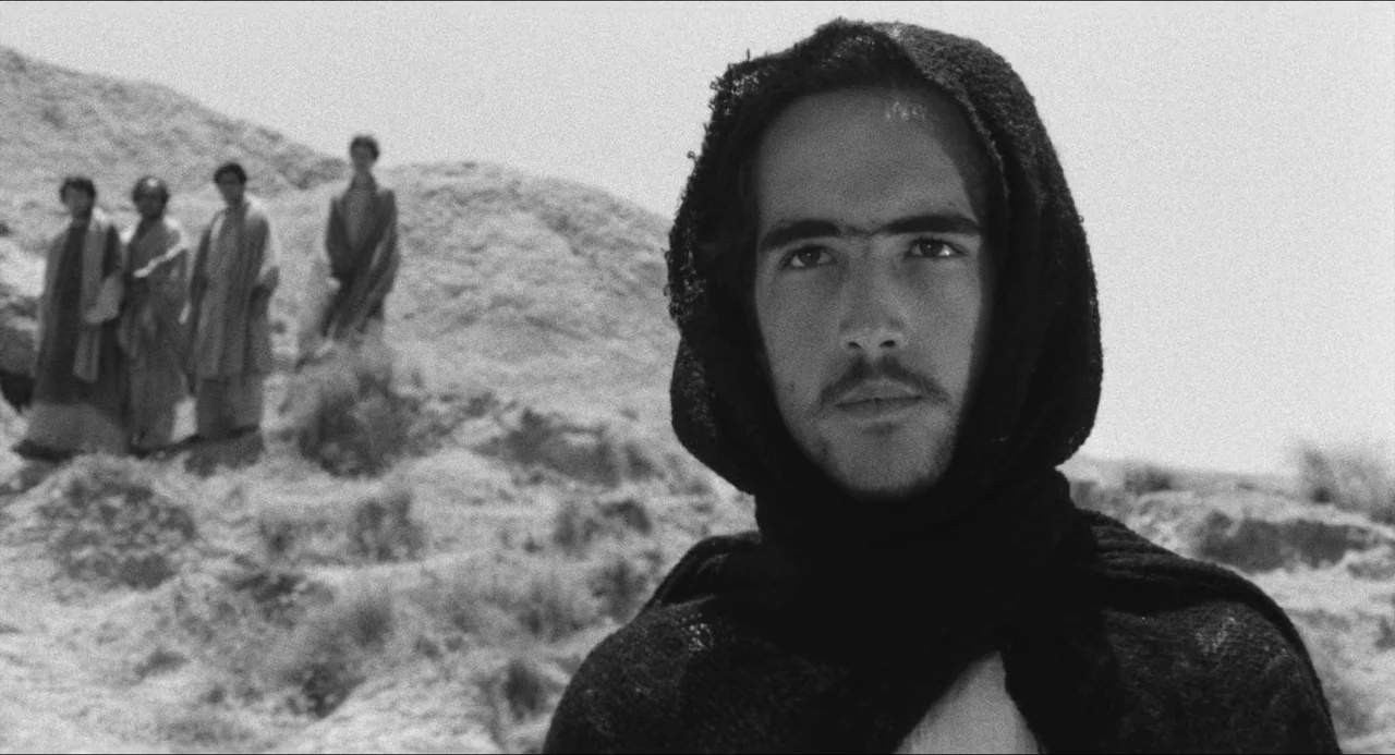 The Gospel According To St. Matthew (1964) movie still: closeup — A dry, rocky desert landscape stretches out behind a group of distant figures. A…