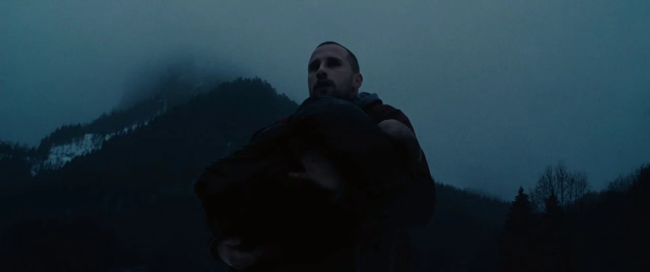Rust And Bone (2012) movie still: medium — A man walks through a dark, foggy mountainous environment during the blue hour. He carries a large, dark,…