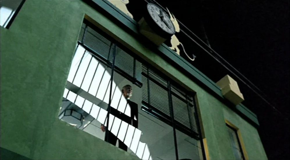 Roselyne and the Lions (1989) movie still: wide — A green concrete building facade features a large clock and reflective windows at night. An…