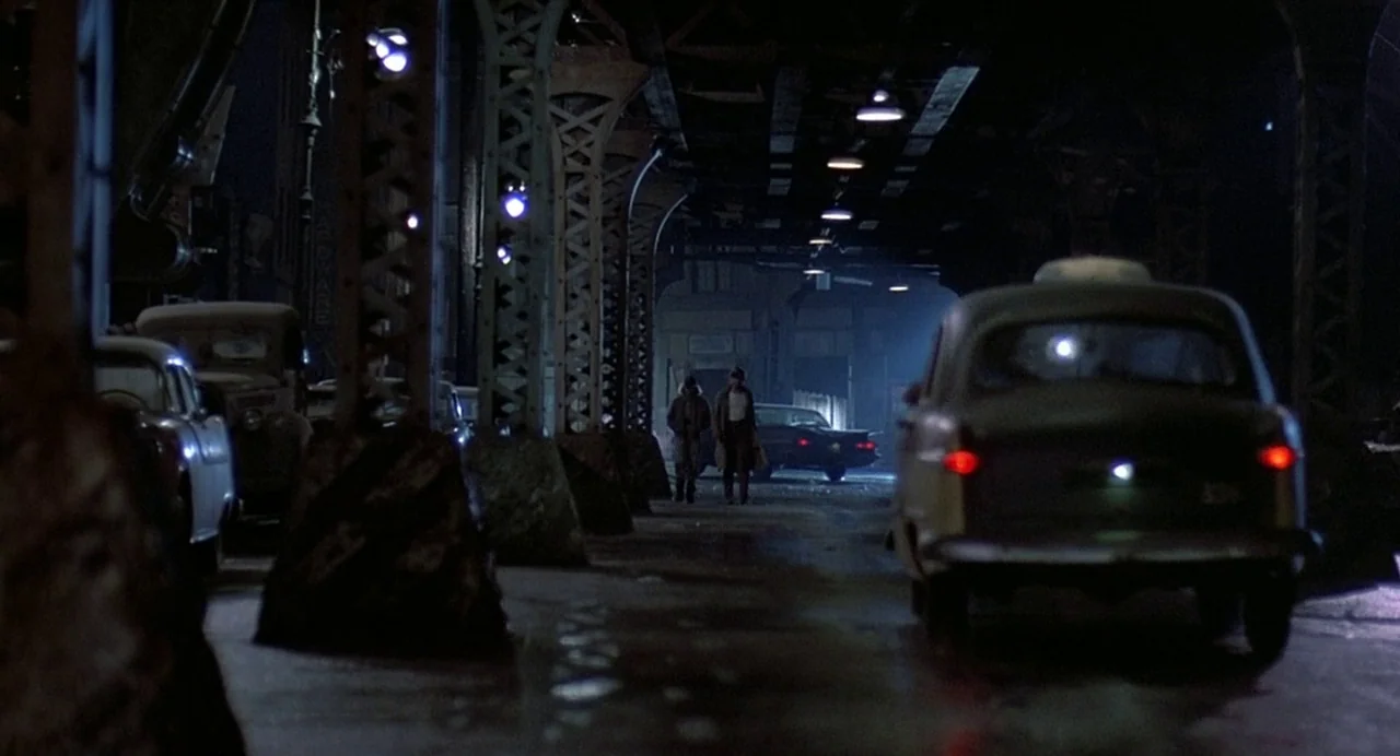 Streets of Fire (1984) movie still: wide — An urban street runs beneath heavy metal support beams of an elevated train line. Two men walk toward the…