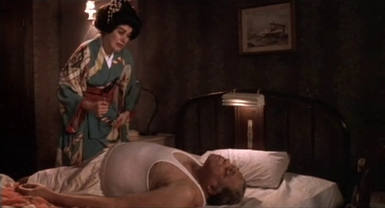 The Annihilation of Fish (1999) movie still: medium — A dimly lit bedroom features floral wallpaper and a framed picture on the wall. A woman in a…