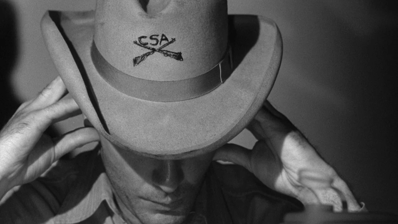 Shock Corridor (1963) movie still: closeup — A man wears a tan cowboy hat with a CSA and rifle emblem on it. The man holds the brim of his hat with…