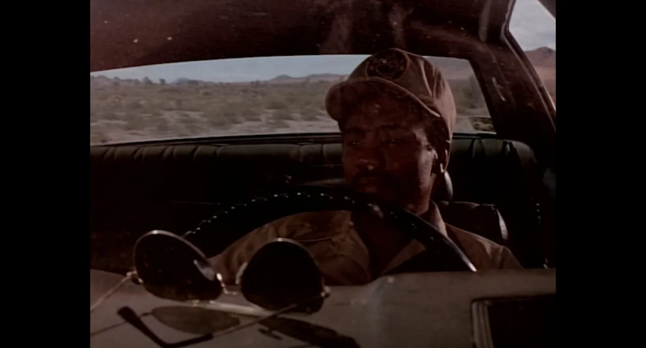 American Yakuza (1993) movie still: medium — A man drives a car through an arid, open desert landscape. He grips the steering wheel while looking…