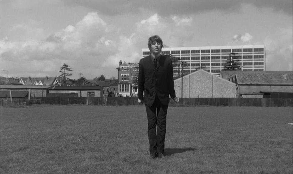 A Hard Days Night (1964) movie still: fullbody — A man stands alone in an expansive grassy field under a cloudy sky. The backdrop features a row of…