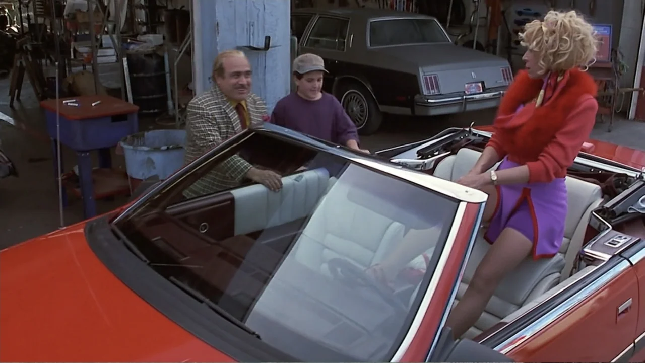 Matilda (1996) movie still: medium — An outdoor auto repair facility features a red convertible car and parked vehicles. A woman standing in the…