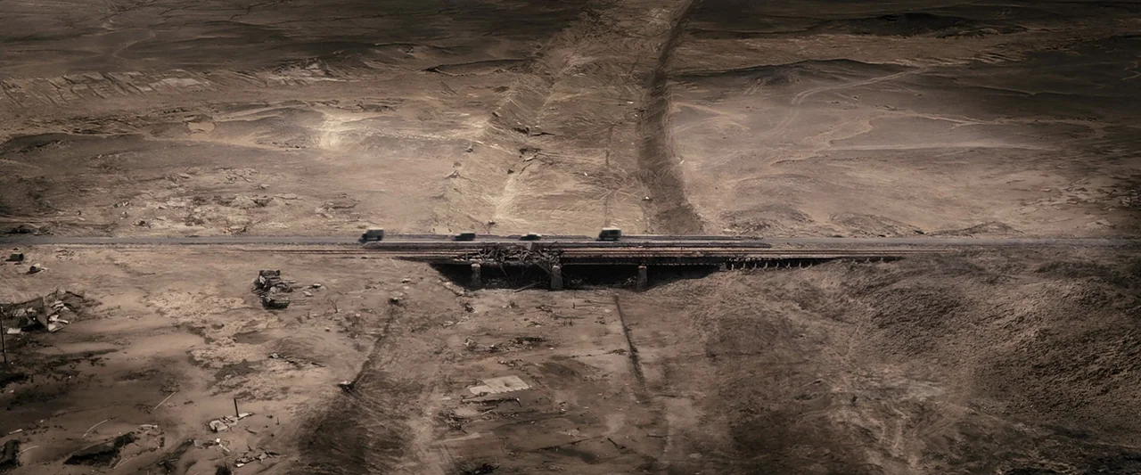 The Book of Eli (2010) movie still: establishing — An arid, desolate landscape features a damaged bridge crossing a dried-up, dusty riverbed. Four…