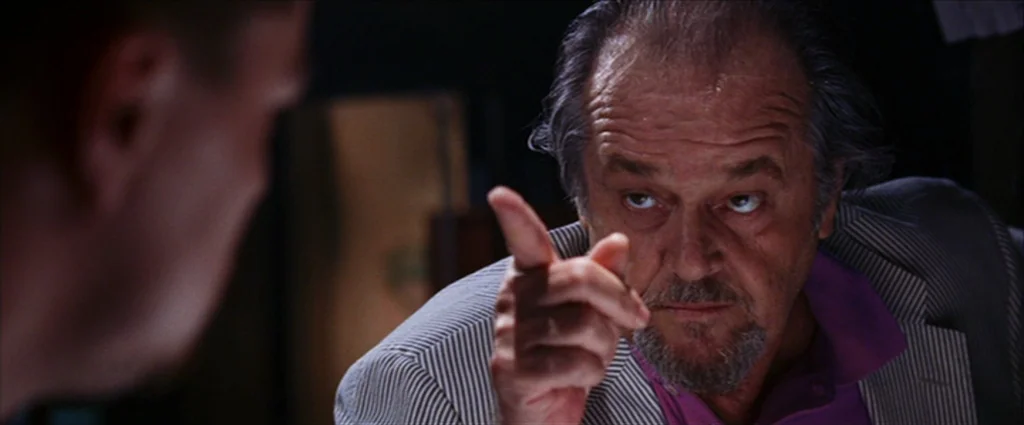 The Departed (2006) movie still: closeup — A man in a striped blazer points a finger while speaking in a dimly lit office. The scene is shot from…