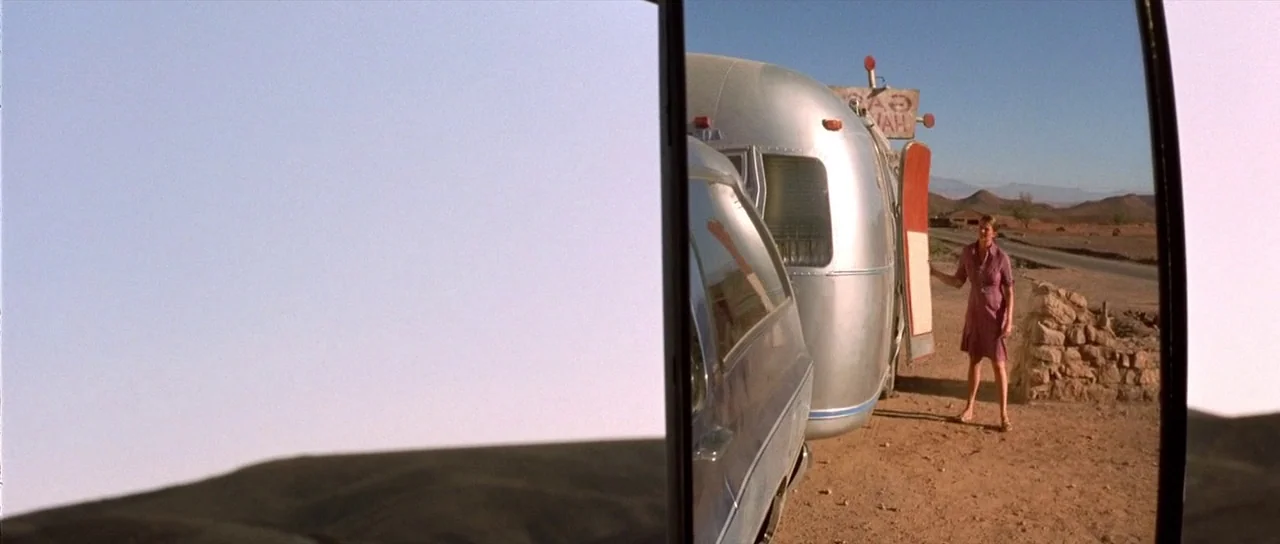 The Hills Have Eyes (2006) movie still: wide — A dry desert roadside features a silver vintage travel trailer parked near a road. A woman in a…