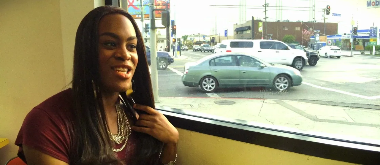 Tangerine (2015) movie still: medium — A view from inside a brightly lit restaurant looking out onto a busy city street. A woman sits in the…