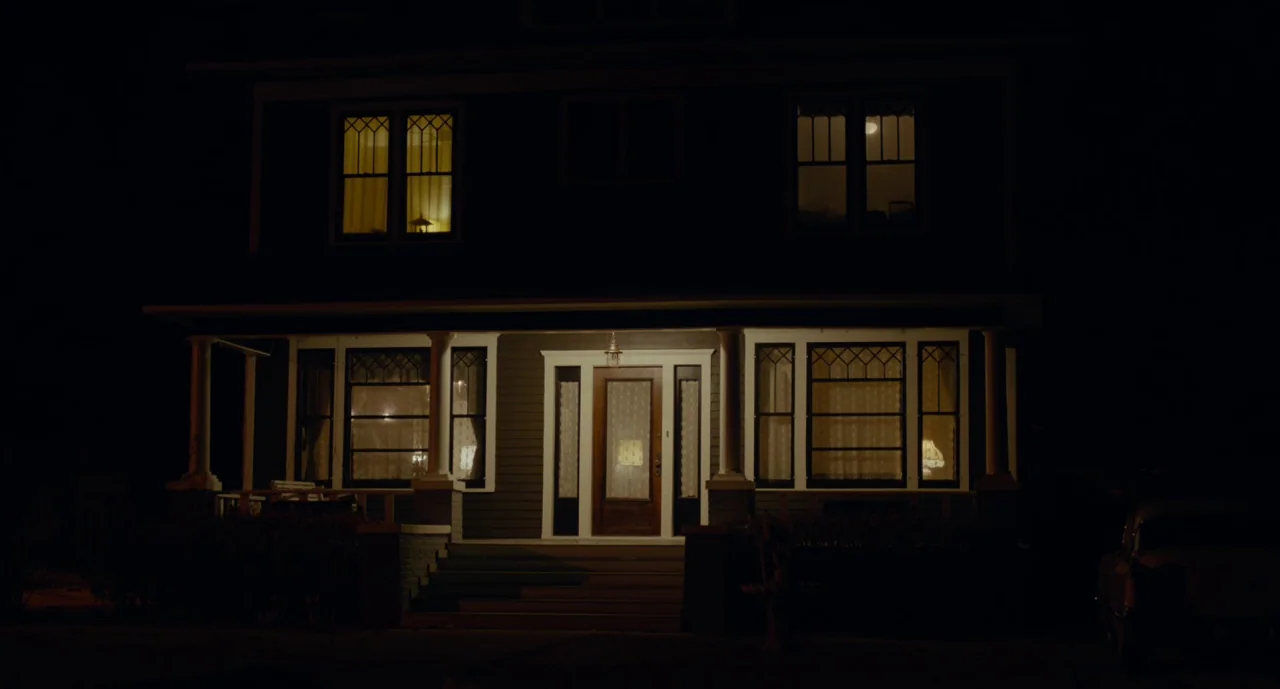 Wildlife (2018) movie still: establishing — A two-story residential home with a covered front porch is illuminated at night. Warm yellow light…