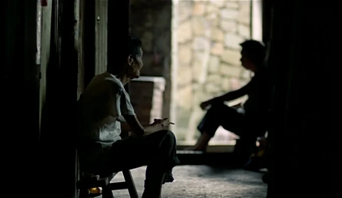 Dust In The Wind (1986) movie still: medium — A dimly lit narrow corridor leads to an exterior stone wall. A man sits smoking on a stool while…