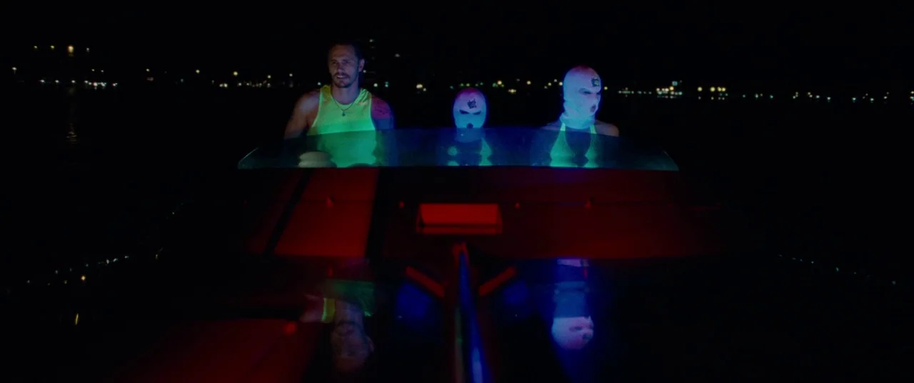 Spring Breakers (2012) movie still: medium — Three individuals sit in a small boat during a dark night on the water. The group wears bright neon…