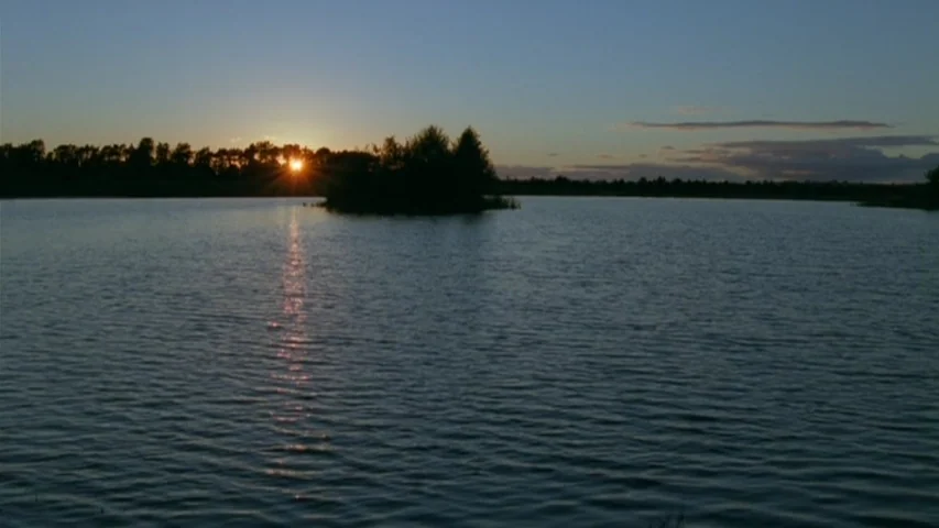 Garage (2007) movie still: establishing — A calm lake stretches toward a distant treeline silhouetted against a setting sun. The light reflects on…