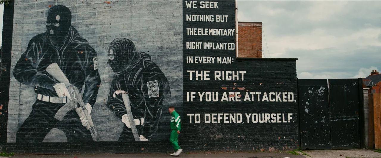 Kneecap (2024) movie still: wide — A brick wall displays a large black-and-white mural of masked paramilitary figures and text. A young man wearing…