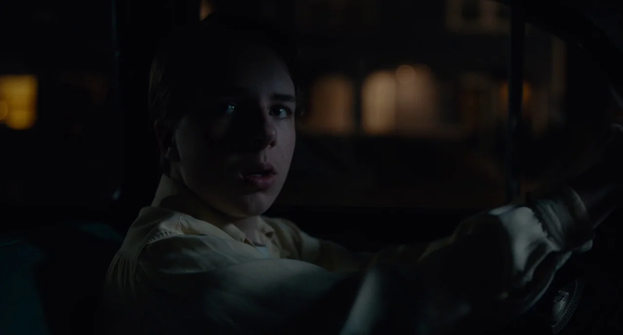 Wildlife (2018) movie still: medium — The interior of a car at night with a teenager at the wheel. The driver looks out the window as the dark…
