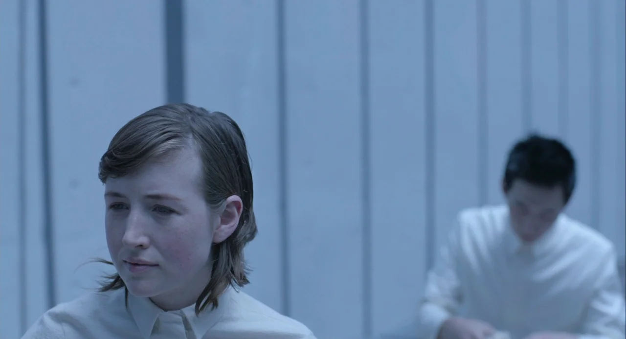 Equals (2015) movie still: medium — A stark white interior with vertical wall panels. Two young individuals sit side by side in matching…