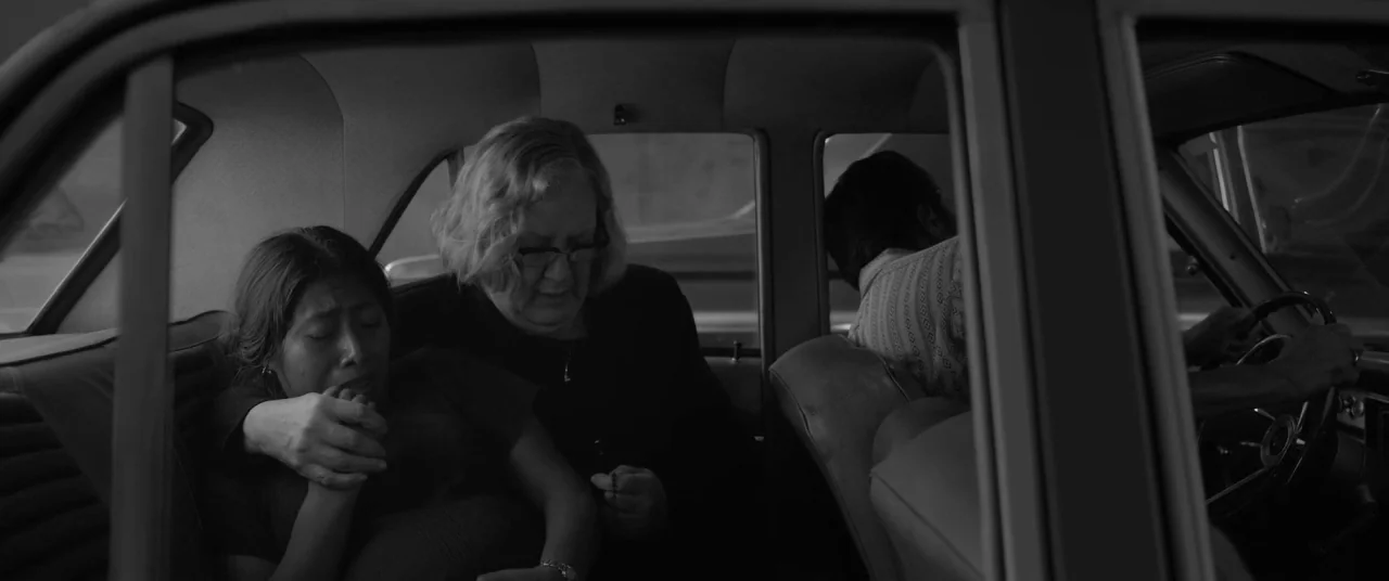 Roma (2018) movie still: medium — The interior of a vintage sedan at eye level shows two women in the back. One woman comfortingly grips the arm of…