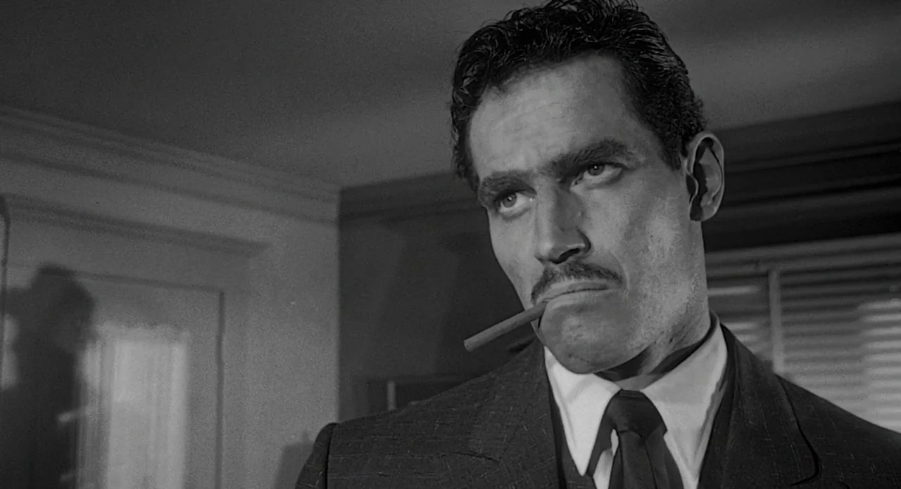 Touch of Evil (1958) movie still: closeup — An interior office with plain walls and horizontal window blinds visible in the background. A man in a…