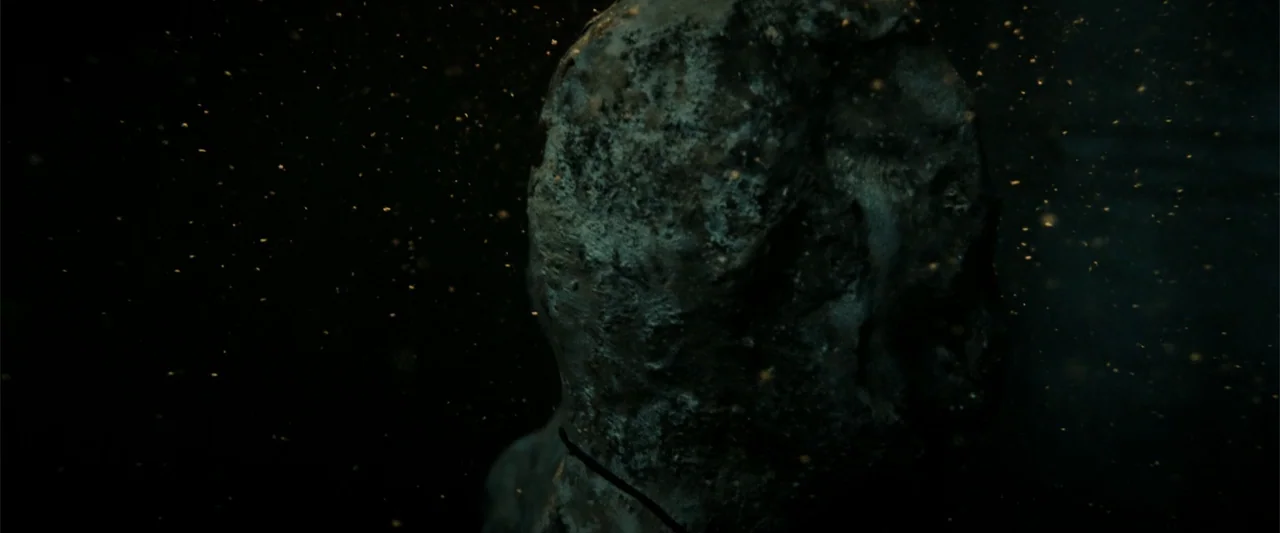 Anaconda (2024) movie still: closeup — A weathered stone bust sits in a dark, atmospheric interior space filled with floating dust. The surface of…