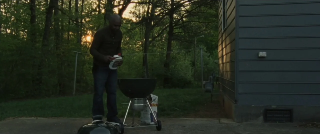 Sermon on the Mount (2019) movie still: fullbody — A residential yard with a grey sided house and thick woods in the background. A man stands on a…