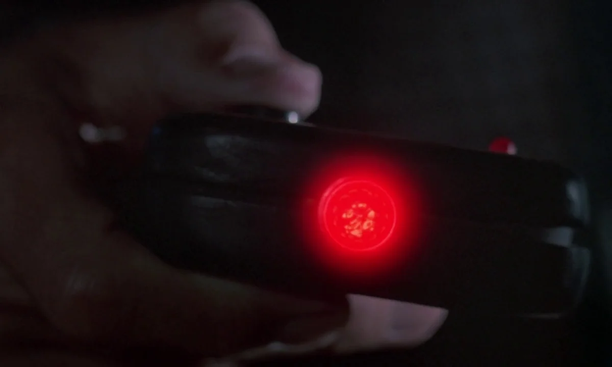 Phenomena (1985) movie still: closeup — A close view of a dark, undetailed area where a black object is held. A small, bright circular red light…