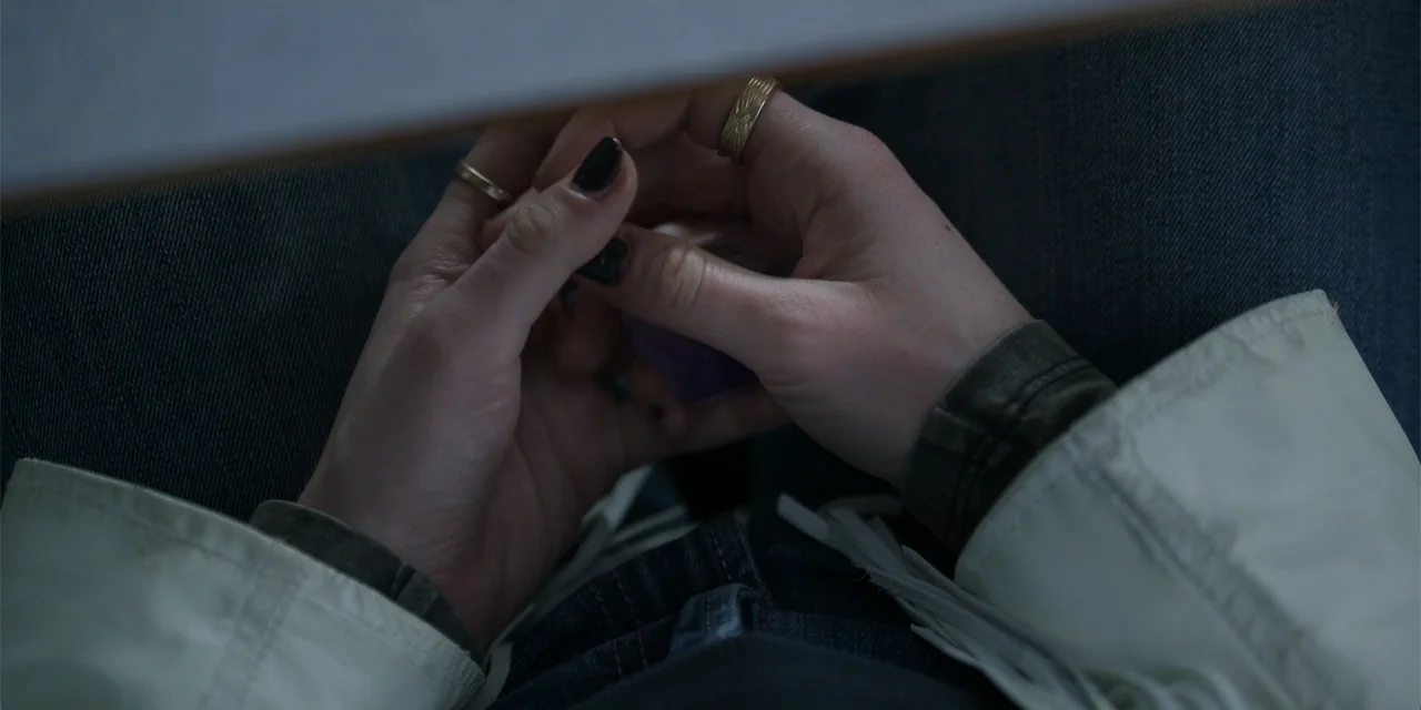 Totally Killer (2023) movie still: closeup — A person sits with hands folded in their lap while wearing jeans and a jacket. Their hands are cupped…