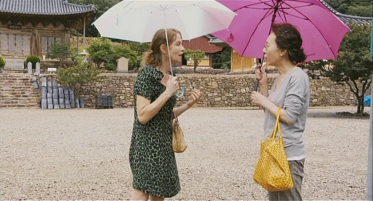In Another Country (2012) movie still: medium — Two women stand on a gravel path in front of traditional wood temple structures. The women converse…