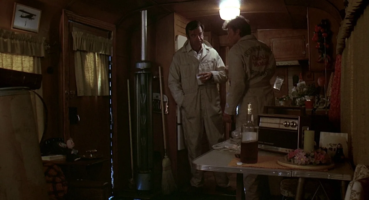Charley Varrick (1973) movie still: medium — A cramped 1970s travel trailer interior features wood paneling and vintage period decor. Two men in…