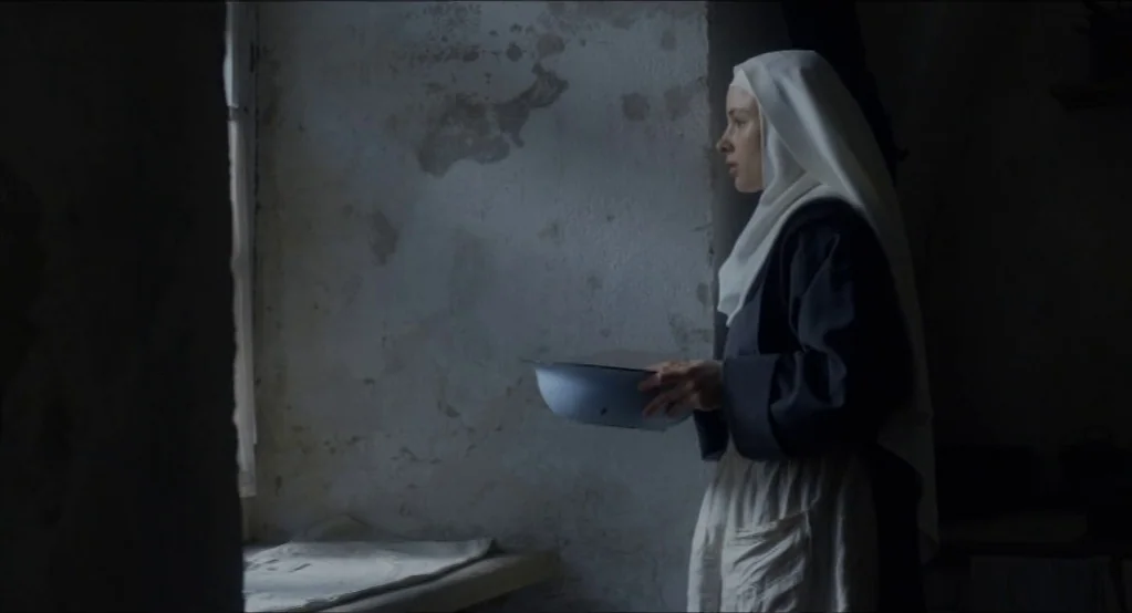 The Innocents (2016) movie still: medium — A stone room with weathered walls and a simple window provides a spare setting. A woman in a nun's habit…