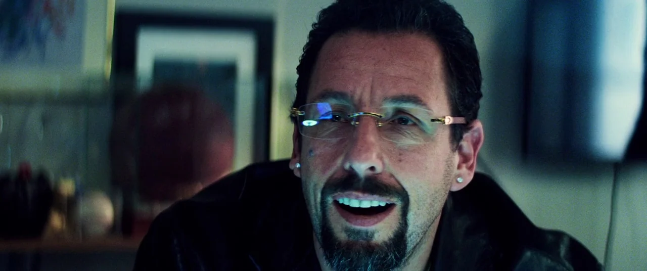 Uncut Gems (2019) movie still: closeup — A dark, cluttered room with framed art and out-of-focus objects in the background. A middle-aged man with a…