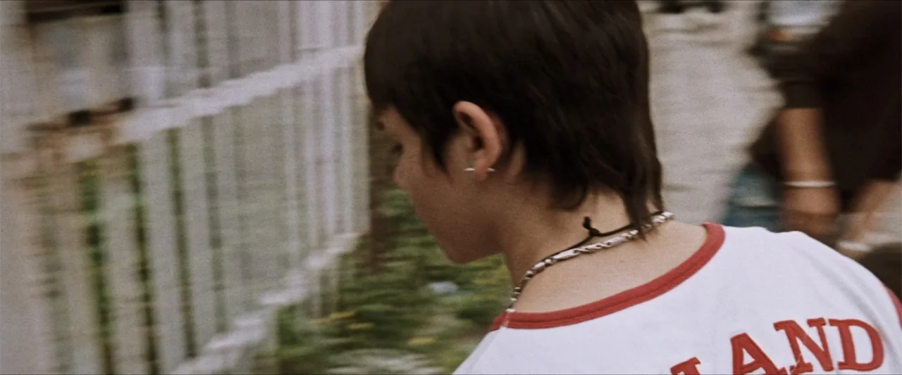 GOMORRAH (2023) movie still: medium — A narrow alleyway features a weathered white fence and blurred concrete wall textures. A teenage girl with…