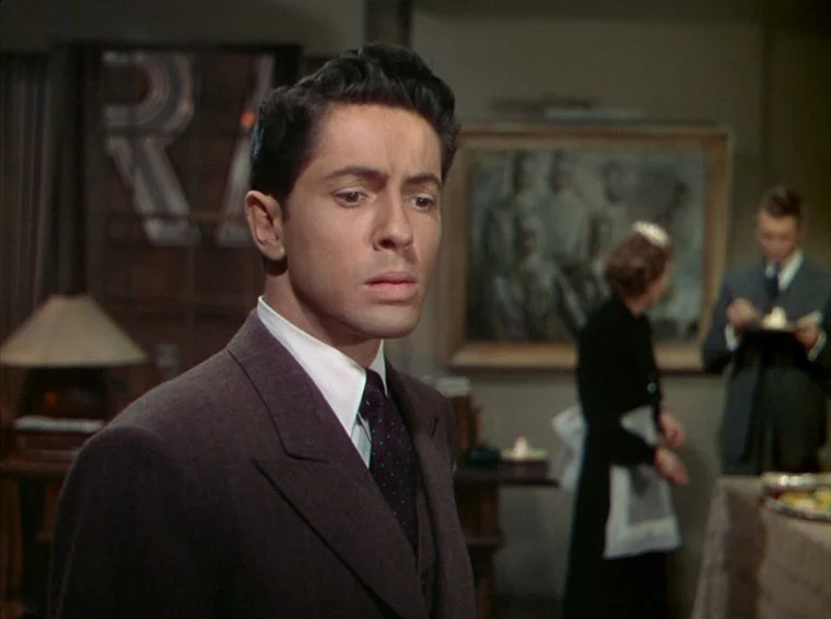 Rope (1948) movie still: medium — A well-furnished room features a painting on the wall and a serving area nearby. A young man in a suit stands in…