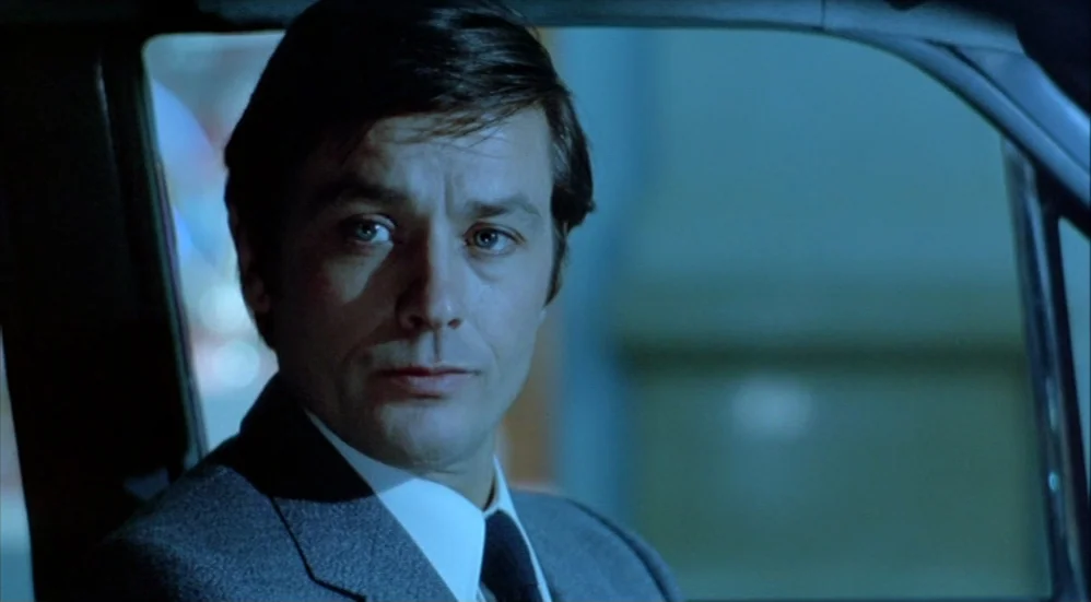 Un Flic (1972) movie still: closeup — A man sits inside a vehicle at night with blurred city lights visible outside. The lighting reflects off the…