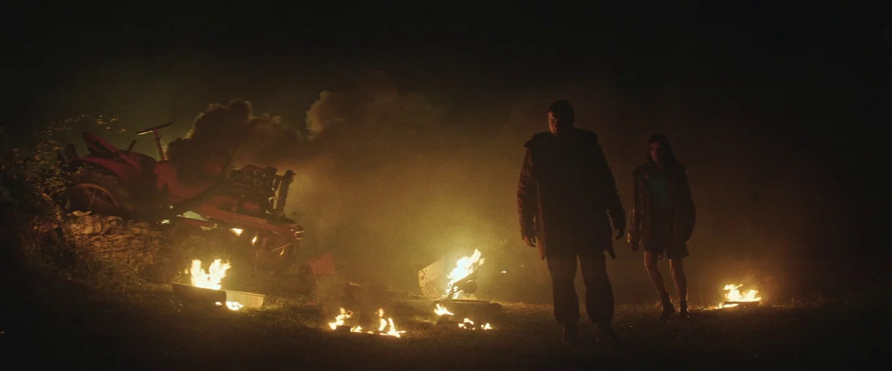 Vincent Must Die (2023) movie still: wide — A dark, grassy field at night with scattered small fires burning on the ground. A man and a woman walk…