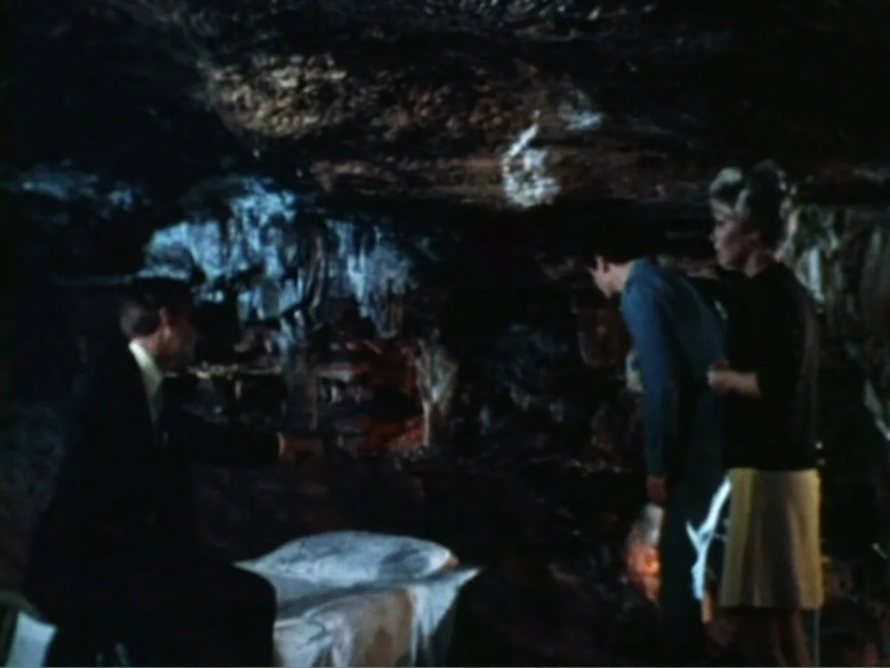 It's Alive (1974) movie still: medium — A dark, cramped interior of a rock cave with stalactite formations on the ceiling. One man sits on a white…