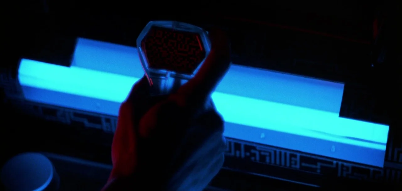 Tron (1982) movie still: closeup — A dark environment features a large blue illuminated panel with intricate geometric patterns. A hand holds a…