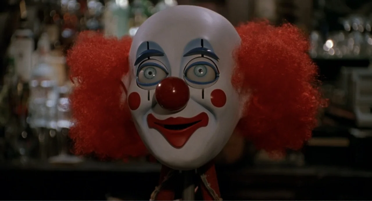 F/X2 (1991) movie still: closeup — A clown mask with bright red hair sits in a dimly lit bar interior. The mask faces directly forward with painted…