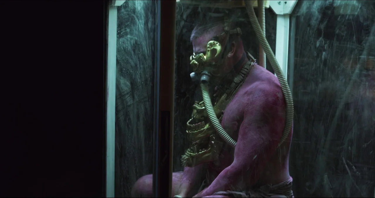 Prospect (2018) movie still: medium — A man with magenta skin sits inside a glass isolation chamber with smudged surfaces. He wears a complex,…