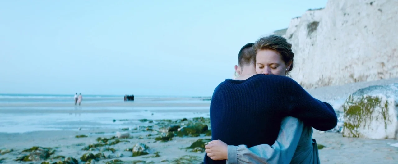 Slack Bay (2016) movie still: medium — A couple embraces on a sandy beach below prominent white chalk cliffs. Distant figures stand on the shoreline…