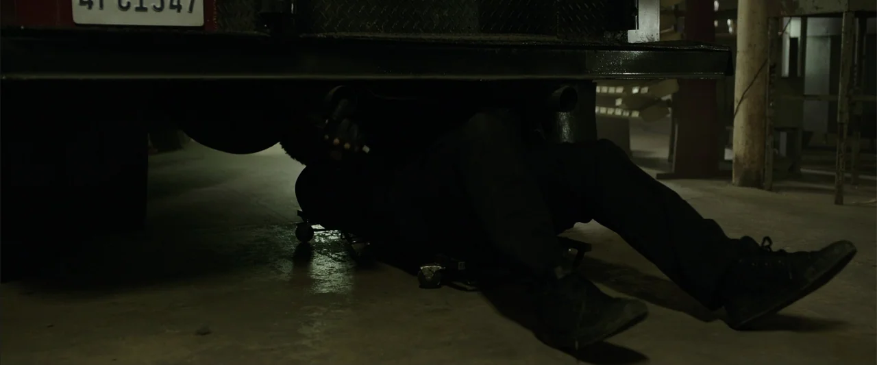 Den of Thieves (2018) movie still: medium — A dimly lit concrete garage floor beneath the rear of a dark vehicle. A person in dark clothing lies on…