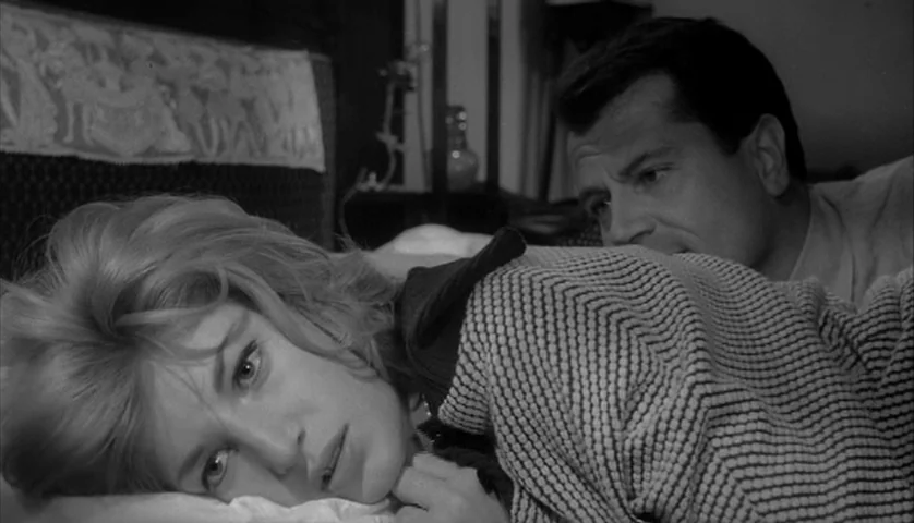 L’Avventura (1960) movie still: medium — A dimly lit bedroom features a bed with textured bedding. A woman looks towards the camera while a man lies…