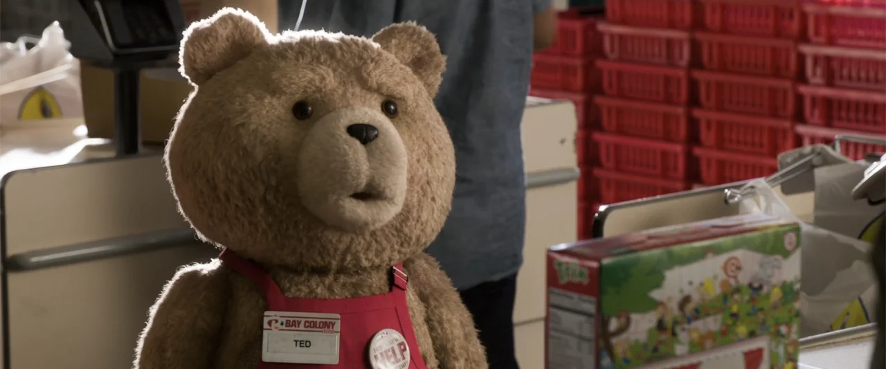 Ted 2 (2015) movie still: medium — A supermarket checkout counter with stacked red plastic crates in the background. A plush teddy bear wears a red…