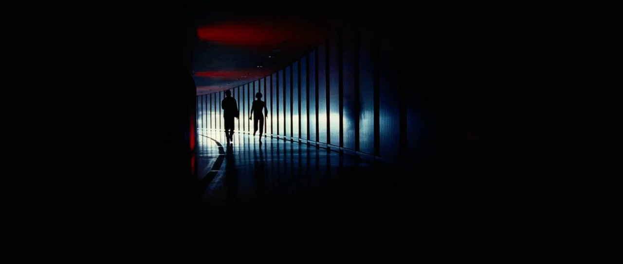 Aeon Flux (2005) movie still: wide — A long, curved modern passageway with a bright backlit wall and dark floor. Two silhouetted figures walk down…