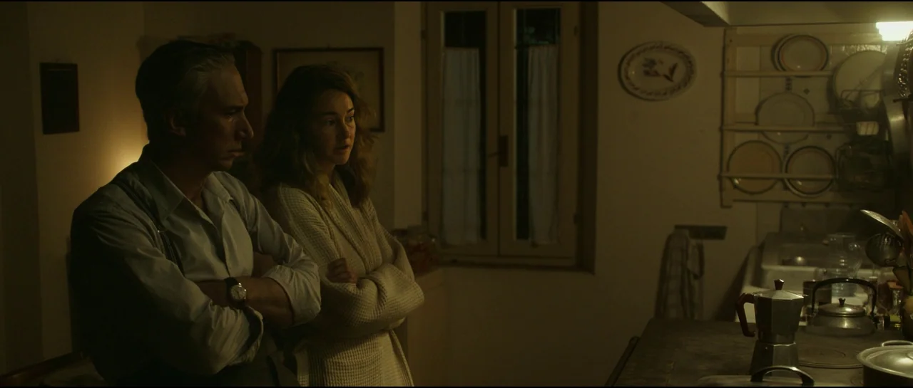 Ferrari (2023) movie still: medium — A dimly lit room with a wooden window, ceramic plates, and an old stove. A man and a woman stand side-by-side…